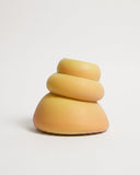 A minimalist, abstract sculpture of three stacked, rounded organic shapes in a warm yellow-orange gradient, set against a seamless light gray background.