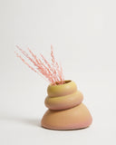 A three-tiered ceramic vase with a smooth, rounded organic shape, featuring a color gradient from pale yellow-green at the top to a warm peachy pink at the base. It is styled with dried pink grass stems and is photographed on a clean, light off-white background.