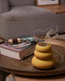 A yellow ceramic vase with a tiered, stacked design sits on a decorative round tray in a cozy living room setting, styled with a book, magnifying glass, and dried grass.