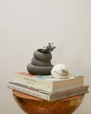 A matte dark gray stacked ceramic planter holding small dried green stems, displayed on top of two books next to a white marble sphere, set on a round wooden side table against a plain off-white wall.
