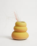 A three-tiered ceramic vase in a matte mustard yellow finish, styled with dried pinkish-tan grass stems, set against a clean white background.
