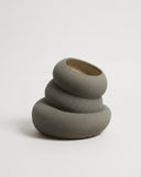 A minimalist, sculptural ceramic planter with three stacked, rounded tiers, finished in a matte dark gray with a contrasting warm beige interior, set against a clean, light gray studio background.