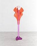 Sculptural Lotus floor lamp featuring layered orange petal-shaped elements, a slender pink stem, and a purple base. Its floral-inspired silhouette combines expressive color with contemporary decorative lighting design.