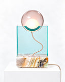 Adrian Cruz Rotonda Onyx Lamp featuring a luminous spherical shade above a transparent turquoise rectangular panel and a richly patterned stone-like base. Its curved metallic detailing and contrasting geometric elements create a distinctive sculptural lighting piece.