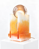 Adrian Cruz Rotonda Sand Orange Lamp featuring a glowing spherical shade paired with translucent orange panels and a warm, layered rectangular base. Its architectural composition and amber-toned palette create a striking contemporary table lamp.