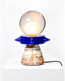 Adrian Cruz Tona Blue Lamp featuring a spherical glass shade, a vivid blue sculptural disc, and a tapered base with warm marbled patterning. Its contrasting geometric shapes and materials create an expressive contemporary lighting accent.