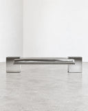 CoExist bench featuring a slim reflective stainless steel seat and solid metallic supports with a clean geometric profile. The polished finish creates a sleek contemporary furniture piece with an architectural silhouette.