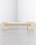 CoExist bench crafted from light travertine, featuring a long rectangular seat and solid stone supports. The porous natural texture and minimalist form highlight the material's understated character.
