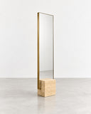 Full-length CoExist mirror featuring a tall reflective panel and a light travertine base with natural porous texture. The understated stone pedestal complements the mirror's slim architectural profile.