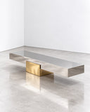 X bench featuring a slim rectangular seat and contrasting geometric metallic supports in warm gold and dark finishes. The architectural composition combines contemporary seating with sculptural material contrast.