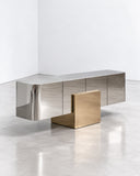 X credenza featuring a long rectangular storage body with reflective metallic surfaces and contrasting geometric gold-toned panels. The clean-lined construction combines contemporary storage functionality with sculptural material contrast.