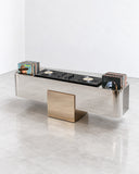 X Deck low storage unit featuring an elongated reflective metallic body with contrasting rectangular gold-toned panels and geometric supports. The architectural design combines functional storage with contemporary sculptural detailing.