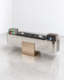 X Deck low storage unit featuring an elongated reflective metallic body with contrasting rectangular gold-toned panels and geometric supports. The architectural design combines functional storage with contemporary sculptural detailing.
