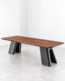 CMX Nashii Dining Table featuring a long, rectangular tabletop in warm-toned wood with visible grain, supported by sculptural dark stone legs. Its bold material contrast and clean architectural lines create a distinctive contemporary dining centrepiece.