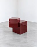 Sculptural CoExist Askew side table featuring a deep red geometric body with offset rectangular volumes and a stepped profile. The bold monolithic design creates a contemporary accent piece with strong architectural character.