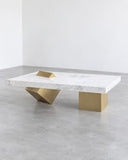 Contemporary CoExist coffee table featuring a pale stone tabletop with subtle natural veining and sculptural geometric brass-tone metal supports. The contrasting materials and asymmetrical base create a refined architectural centerpiece for luxury residential and hospitality interiors.