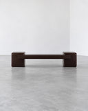 CoExist Redux bench featuring a long, dark ebony-toned seat supported by substantial rectangular end blocks. The minimalist silhouette emphasizes geometric form and understated contemporary design.
