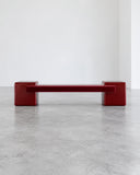 CoExist Redux bench in a rich oxblood red finish, featuring a long rectangular seat and solid geometric end supports. Its bold monochromatic form combines functional seating with sculptural contemporary design.