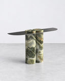 ETAMORPH Jumbo Console features a narrow, elongated dark tabletop balanced on a sculptural cylindrical pedestal in green stone with golden and cream veining. Its asymmetric proportions and richly patterned base give the console a distinctive contemporary presence.