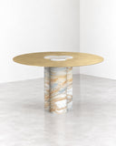 ETAMORPH Jumbo Dining Table features a circular gold-toned tabletop supported by a cylindrical stone pedestal with warm veining. Its clean, sculptural form and contrasting materials bring understated elegance to contemporary dining spaces.