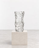IAMMI Melty Size M is a transparent glass decorative object with an irregular, rippled surface and organically distorted cylindrical form. Its light-catching texture creates a sculptural accent for contemporary interiors.