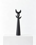 Pich wood sculpture featuring a compact abstract figure with a rounded central form, slender legs, and a small crescent-shaped top. The dark finish and clean sculptural geometry create a distinctive contemporary object.