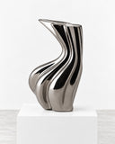 Niccolo Poggi Sinuo Platino Vase featuring a reflective platinum-tone finish with flowing, ribbon-like contours and an asymmetrical flared opening. Its metallic sheen and sculptural silhouette add a refined contemporary accent to luxury interiors.