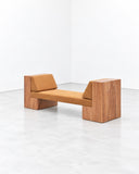 Noemi Saga Ini Wood Daybed featuring a long caramel-toned upholstered cushion framed by substantial wooden end supports with visible grain. Its low, linear profile and warm material palette suit contemporary residential lounges and hospitality spaces.