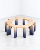 Onno Adriaanse Dawn Circle Bench featuring a circular open-frame wooden seat supported by multiple rounded legs that transition from natural wood to deep blue tones. Its sculptural construction and gradient finish create a distinctive contemporary seating piece.