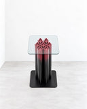 Paolo Giordano Jaipur Garden Side Table featuring a transparent square glass top supported by a glossy black pedestal with sculptural red detailing. Its contrasting materials and decorative central feature create a distinctive contemporary accent table.