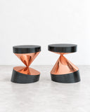 Paolo Giordano Whirling Twins Side Table featuring two sculptural hourglass-shaped bases in a reflective copper finish, topped with round black surfaces. The contrasting metallic curves and dark tops create a bold contemporary design statement.