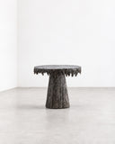 RIES Pasto Dining Table featuring a round tabletop and substantial tapered pedestal base with a rugged, bark-like textured finish in dark earthy tones. Its monolithic silhouette and tactile surface create a distinctive sculptural centrepiece for contemporary dining interiors.