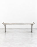 RIES Tori Bench featuring an elongated, narrow seat with subtly upturned ends and slender sculptural legs in a pale grey, stone-like finish. Its restrained linear silhouette and textured surface suit contemporary residential and hospitality interiors.