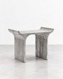 RIES Tori Stool featuring a gently curved seat and two broad, arch-shaped supports in a pale grey, stone-like finish. Its sculptural open base and understated geometric form make it a distinctive contemporary accent seat.