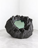 Richard Yasmine Silent Hollows Medium Wall Mirror featuring an organically shaped reflective surface framed by thick, folded black forms. Its rugged sculptural outline and contrasting reflective centre add depth and texture to contemporary interiors.
