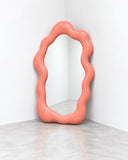 Ryan Mercer Portal Floor Mirror featuring a tall reflective surface framed by an irregular, undulating coral-pink border. Its organic silhouette creates a sculptural focal point for contemporary residential and hospitality interiors.