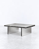 Arris coffee table featuring a slim rectangular reflective metal top and a geometric metallic base. Its low profile and precise lines combine functional surface space with contemporary architectural design.