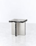 Arris side table featuring a compact reflective metal tabletop and a sculptural rectangular pedestal base. The polished finish and minimalist geometry create a refined contemporary accent piece.