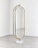 Giove floor mirror featuring a tall oval reflective panel framed by a slender gold-toned metal structure with an elongated arched outline. The minimalist design combines full-length functionality with sculptural contemporary detailing.