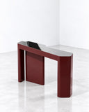 Kara console table featuring a bold burgundy-red rectangular top and matching sculptural pedestal supports with angular cutouts. Its monolithic silhouette and saturated finish create a striking contemporary furniture piece.