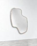 Lab L wall mirror featuring an elongated irregular reflective surface outlined by a slim light-toned frame. Its organic asymmetrical silhouette creates a contemporary architectural accent.