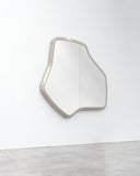 Lab M wall mirror featuring a compact freeform reflective surface and a slim light-toned frame with softly angled contours. Its asymmetrical geometry adds sculptural character to contemporary interiors.