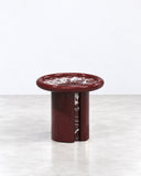 Moonrise side table featuring a circular burgundy stone top with expressive pale veining and a cylindrical pedestal base. The compact sculptural form highlights the natural variation of the stone.