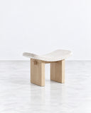 Nara stool featuring an organically shaped pale seat supported by two solid light-toned wooden legs with rounded edges. The natural timber and fluid seat profile create a refined contemporary accent.