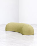 Nuba bench featuring an elongated, softly curved upholstered seat in muted lime-green fabric. Its low, continuous silhouette creates a sculptural contemporary seating piece with a tactile finish.