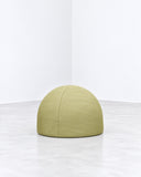 Nuba pouf featuring a compact rounded upholstered form in muted lime-green fabric with a softly curved profile. Its minimalist sculptural shape offers versatile contemporary seating.
