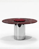 Sculptural ORBITAE dining table featuring a richly veined burgundy stone tabletop supported by a cylindrical polished metal base. The contrasting materials create a refined contemporary centerpiece for sophisticated dining spaces.
