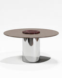 Contemporary ORBITAE dining table with a circular burgundy stone tabletop and reflective cylindrical metal pedestal. Its sculptural construction combines natural stone character with sleek architectural detailing.