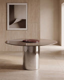 ORBITAE round dining table featuring a burgundy veined stone top and cylindrical metallic base. The sculptural silhouette balances the organic variation of natural stone with refined contemporary geometry.