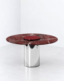 Orbitae dining table featuring a round burgundy stone tabletop with natural veining and a reflective cylindrical metal pedestal. Its sculptural proportions combine natural stone character with sleek contemporary detailing.
